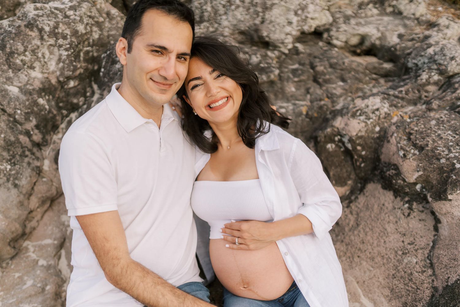 Happy couple sitting against rocks, woman showing pregnant belly, both smiling warmly in white outfits.