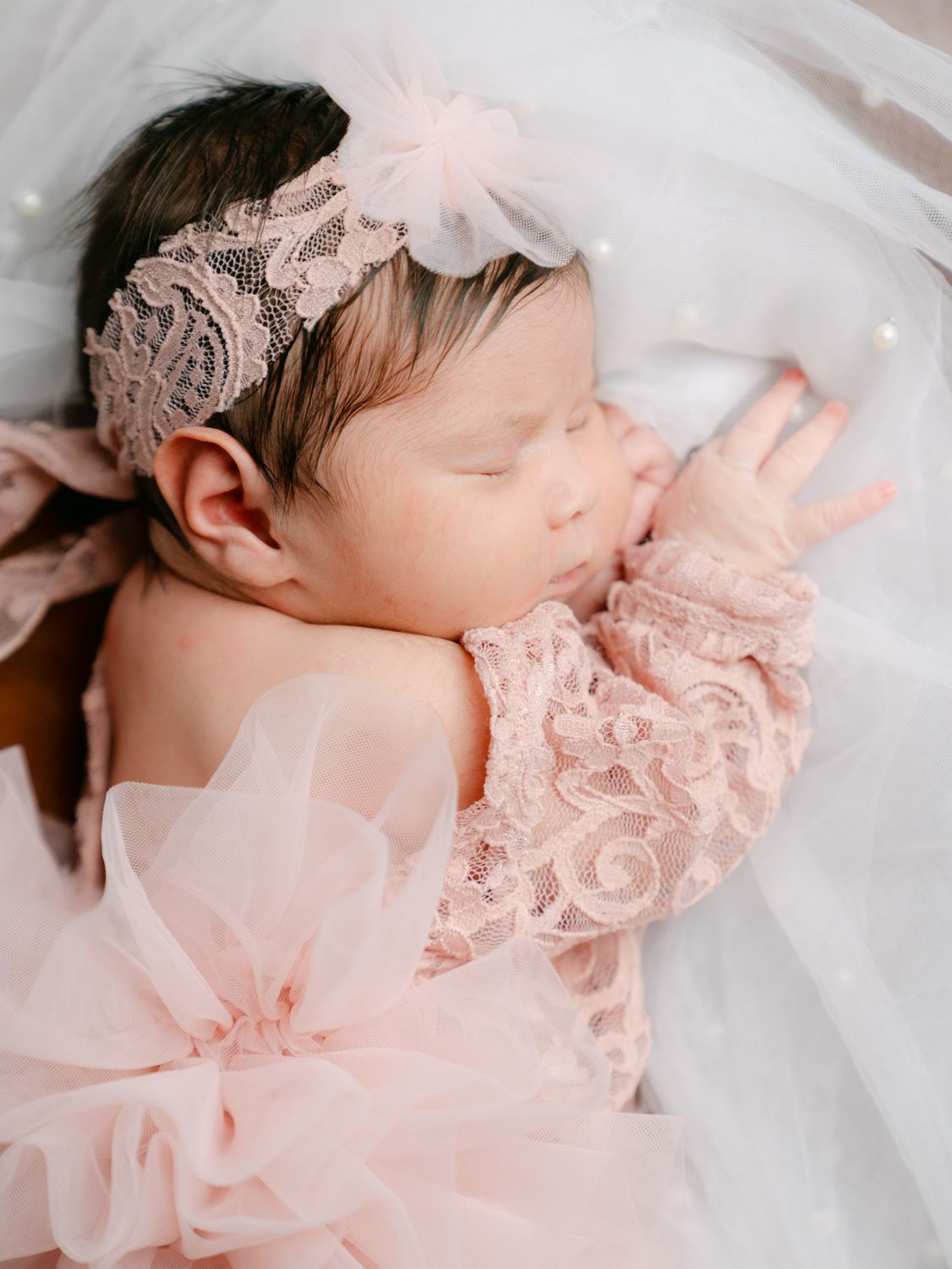 Sleeping newborn baby in pink lace dress and floral headband, peacefully resting on white tulle fabric.