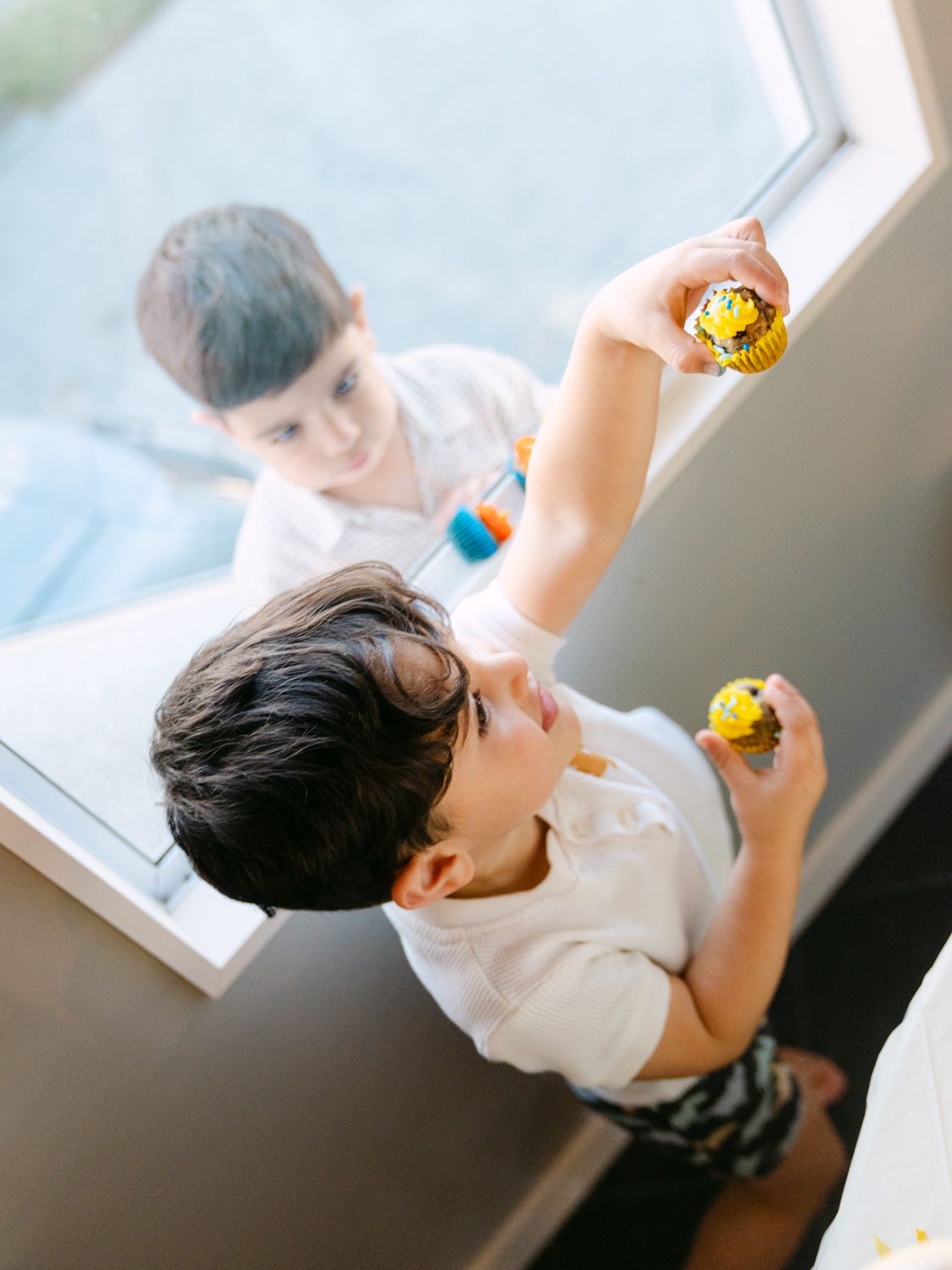 Two young boys playing with small colorful toys near a bright window indoors.