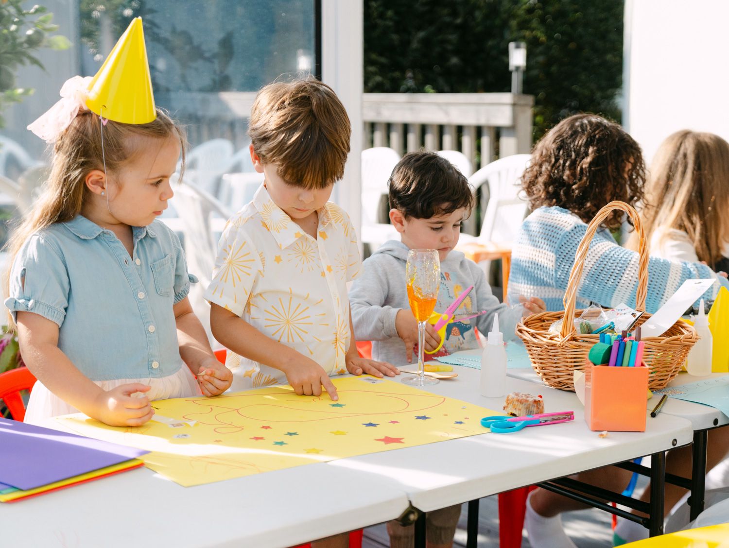 Children doing crafts at a birthday party table with colorful paper, markers, and a wicker basket of supplies.