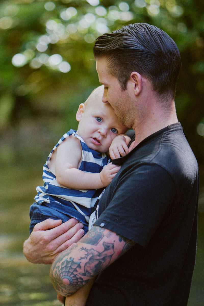 Tattooed father tenderly holding his baby in blue striped romper outdoors in a lush green setting.