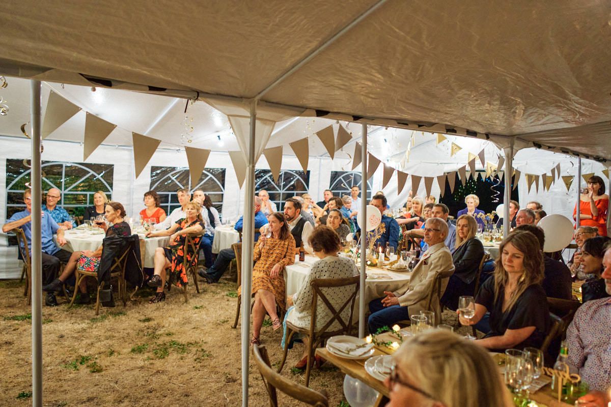 Guests dining under a festive white tent with bunting lights at an outdoor evening party event.