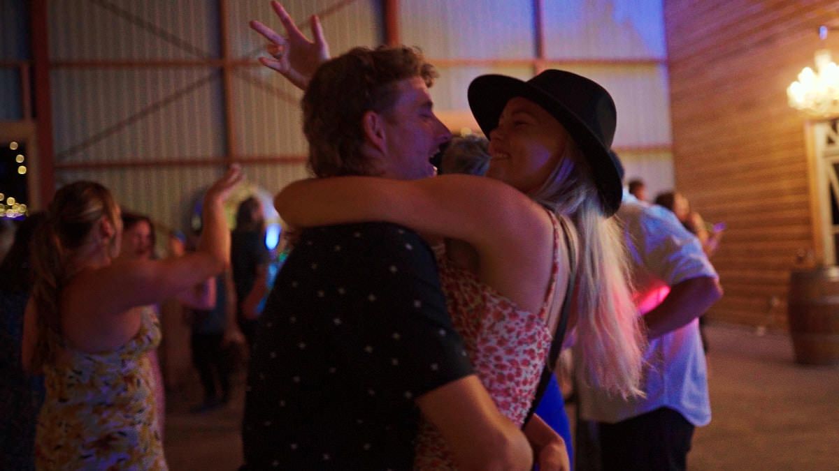Couples dancing closely at a lively barn party with other guests celebrating in the background.