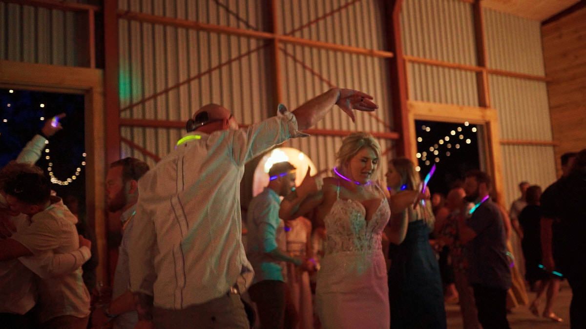 Wedding guests dancing joyfully at a barn reception with glowing neon sticks and string lights.