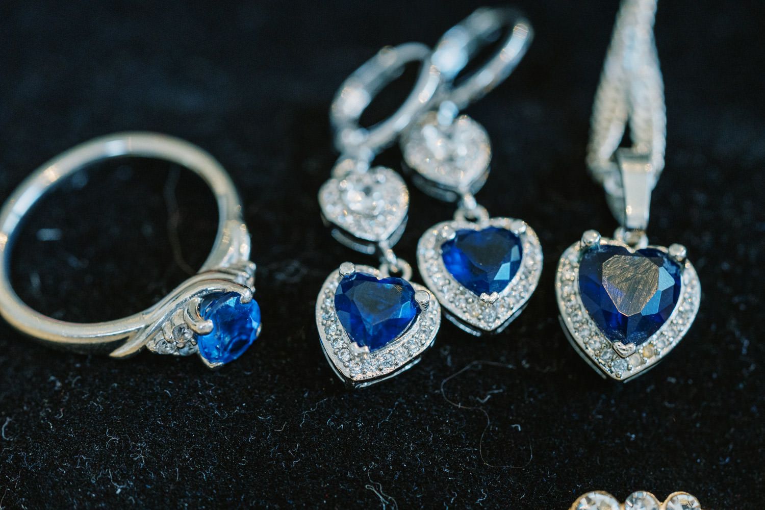 Blue sapphire heart-shaped earrings and ring set with diamond halo accents in silver on dark velvet background.