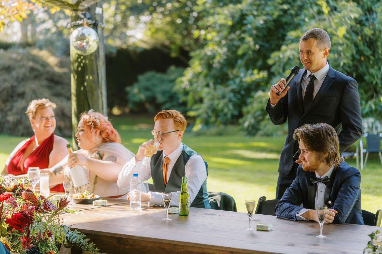 Wedding guests gathered at an outdoor reception table while a man in a black suit gives a heartfelt toast.