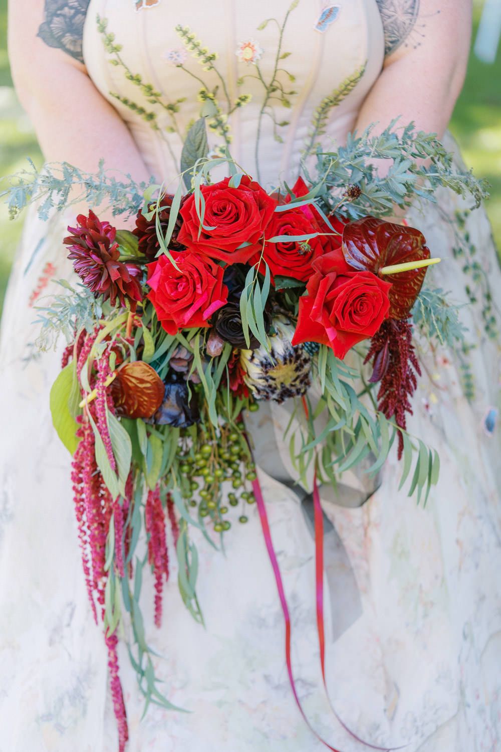 Bride holding a lush red rose bouquet with cascading greenery and burgundy accents in a floral wedding dress.