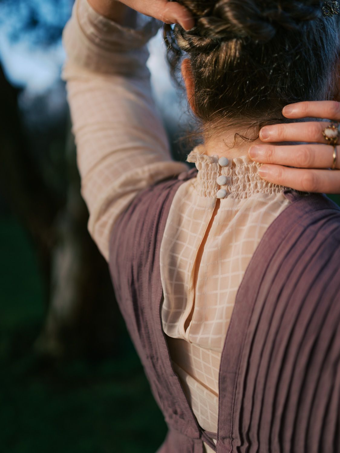 Woman in vintage mauve dress with lace collar, hands touching her upswept hair, photographed from behind outdoors.