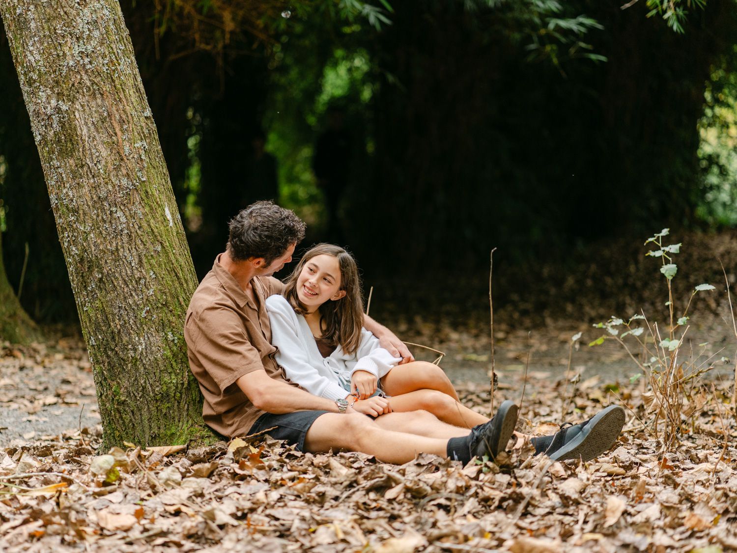 Couple sitting together against a tree trunk on autumn leaves in a lush green forest, smiling and enjoying each other.