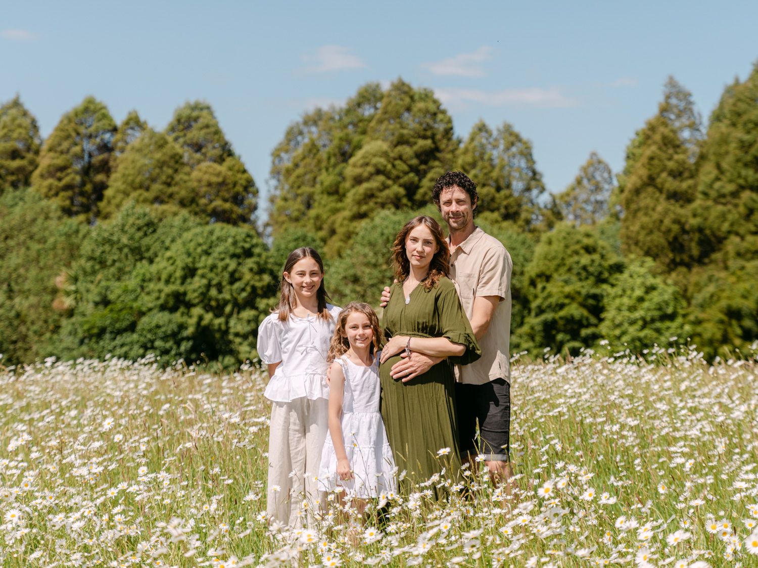 Family of four posing in a white wildflower field with tall green trees under a bright blue sky.