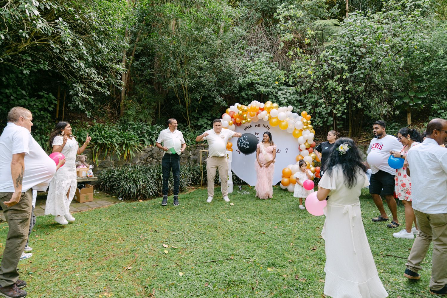 Outdoor gender reveal party with balloon arch in gold and white, guests gathered on green lawn surrounded by lush trees.