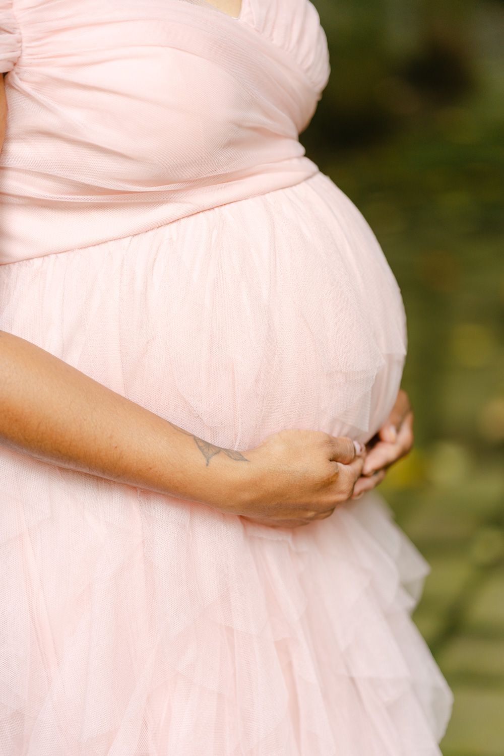 Pregnant woman in pink tulle dress cradling baby bump outdoors, maternity photography session.