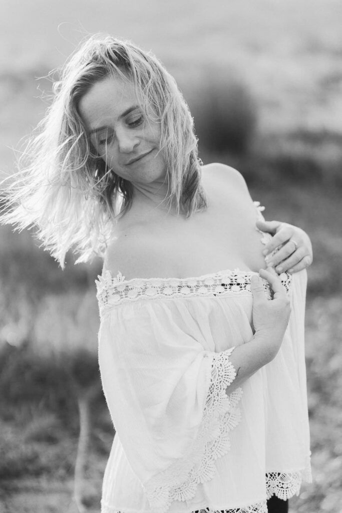Black and white photo of a blonde woman in an off-shoulder white lace dress, standing outdoors with wind-blown hair.