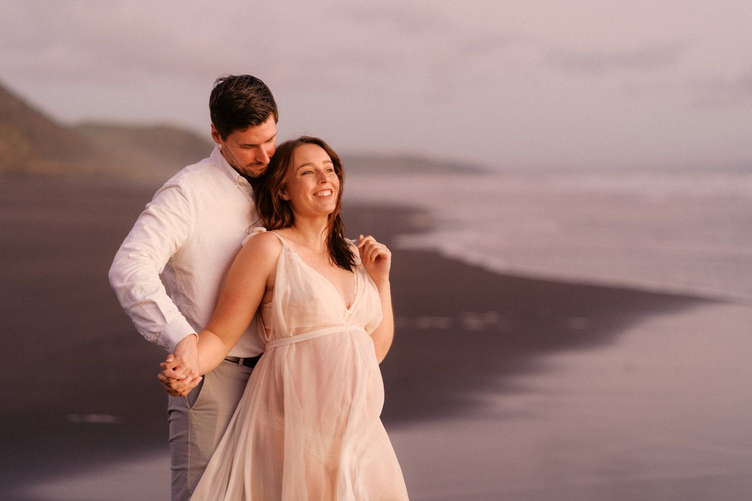 Couple embracing on black sand beach at sunset, woman in sheer dress smiling, man in white shirt holding her hands.