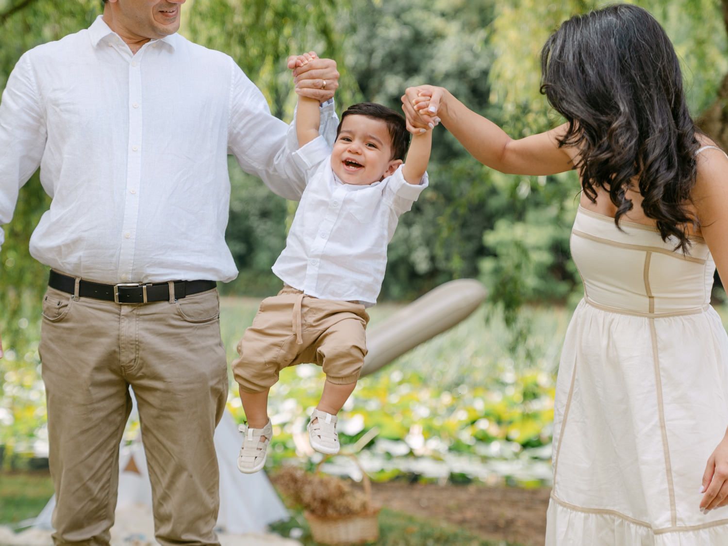 Happy toddler being swung by parents in a lush green park, family portrait in neutral tones.