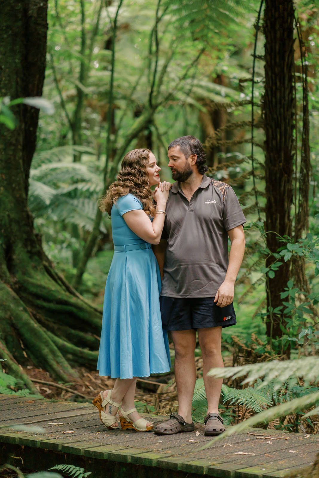 Couple sharing a romantic moment on a wooden bridge in a lush green forest, woman in blue dress.