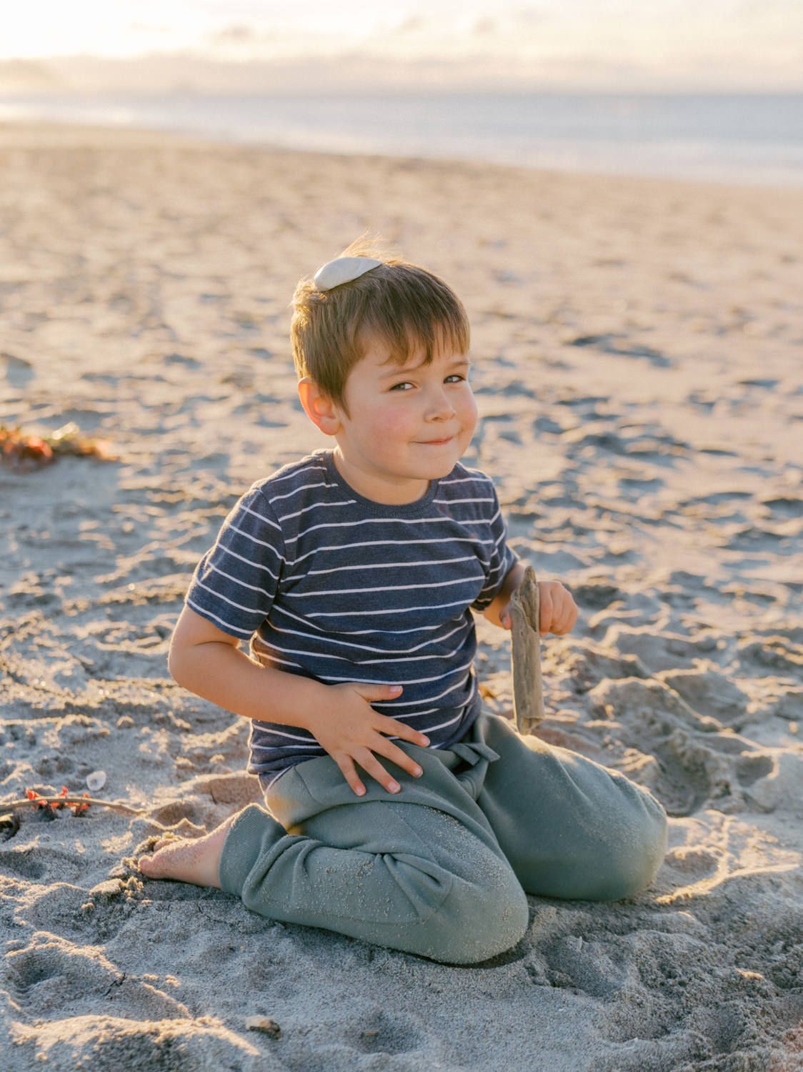 A young boy in a striped shirt sits cross-legged on a sandy beach at golden hour, holding a stick.