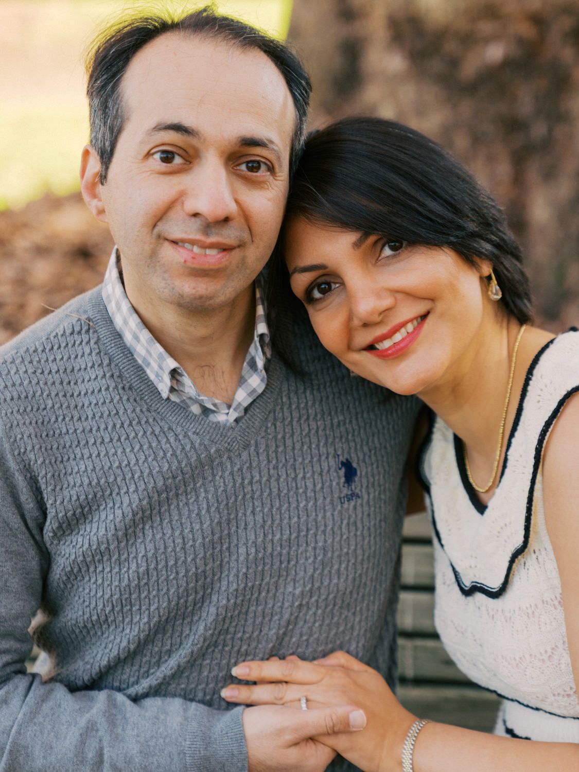 A smiling couple poses closely together outdoors, the woman resting her head on the man's shoulder affectionately.