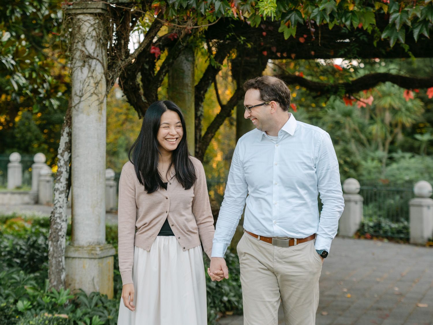 Couple holding hands and smiling while walking through a lush garden pathway with stone pillars.
