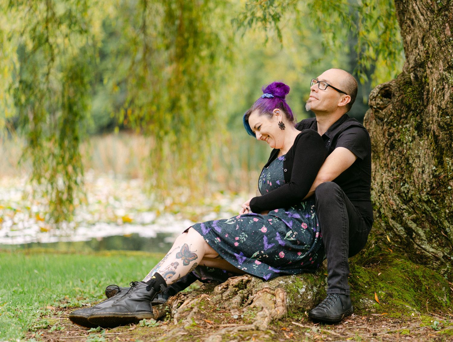 Couple with purple hair and tattoos sitting together under a willow tree in a lush green park setting.