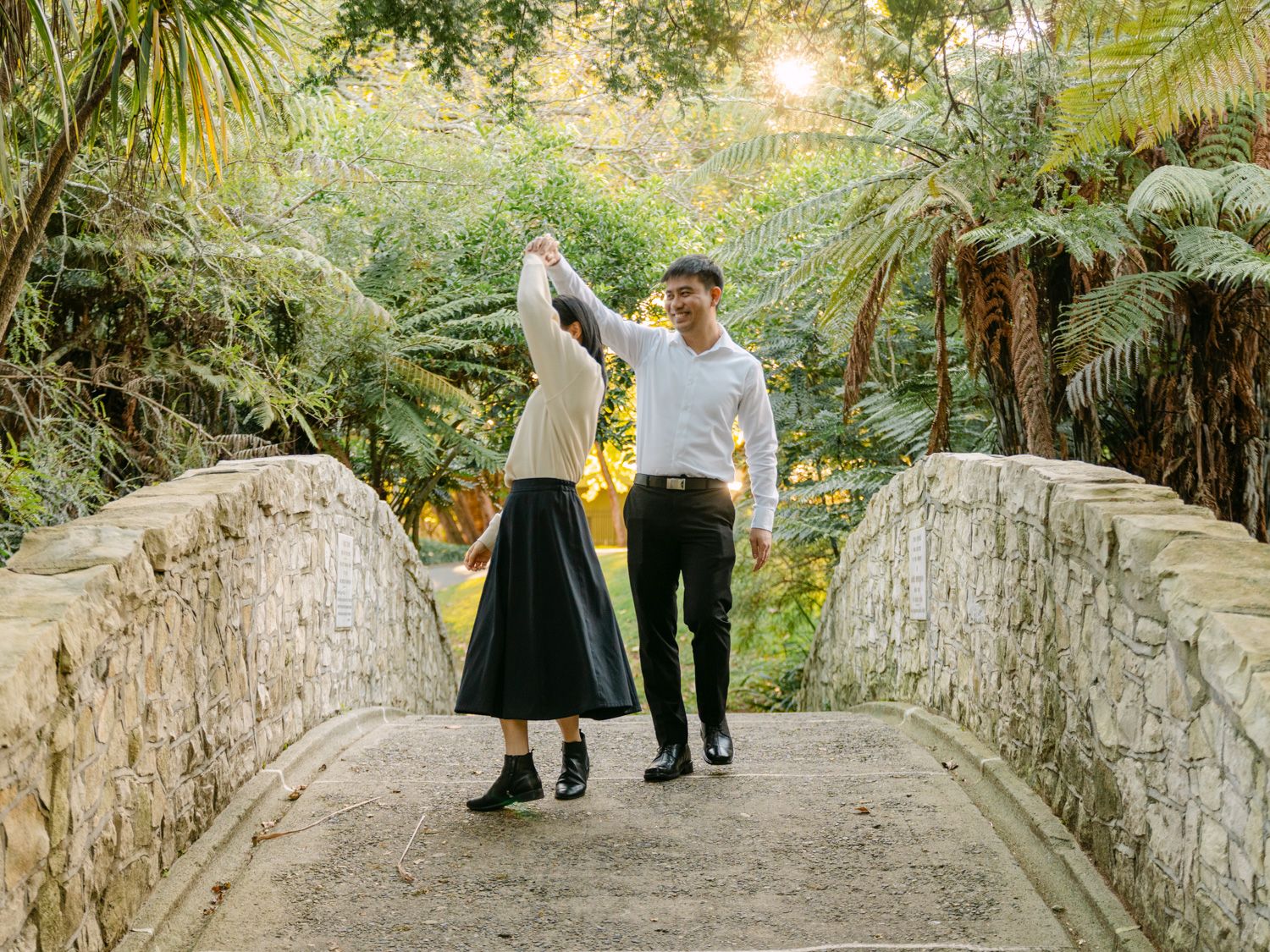 Couple dancing on a stone bridge surrounded by lush tropical greenery and palm trees in a scenic garden.