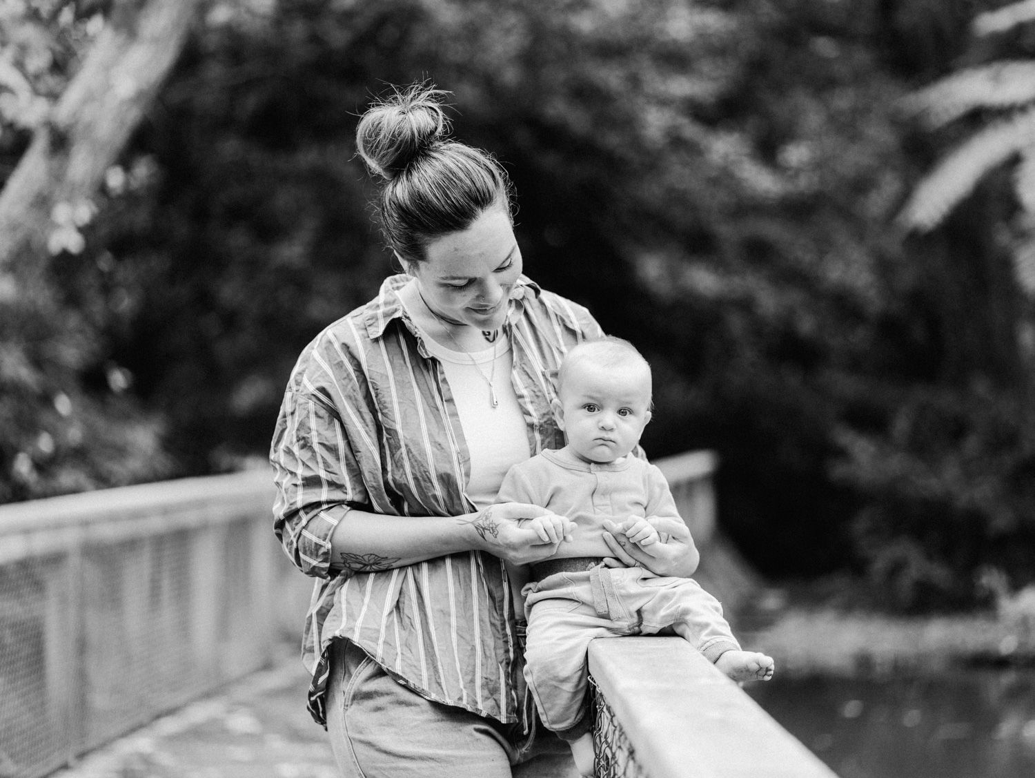 Black and white photo of a mother holding her baby on a wooden bridge surrounded by lush greenery.