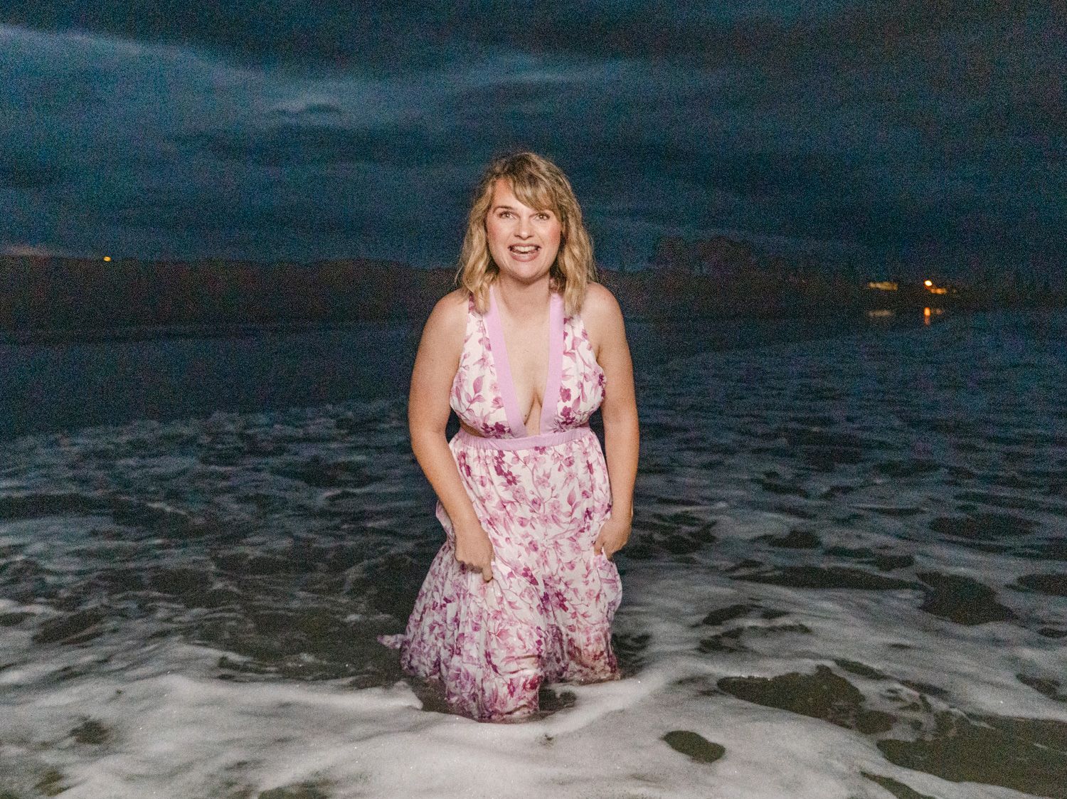 Woman in floral dress standing in ocean waves at dusk with dramatic cloudy sky.