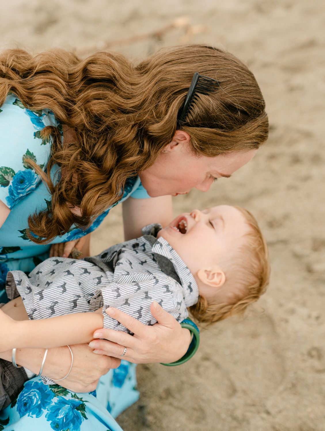 A mother lovingly holds her laughing toddler on a sandy beach, both wearing blue outfits.