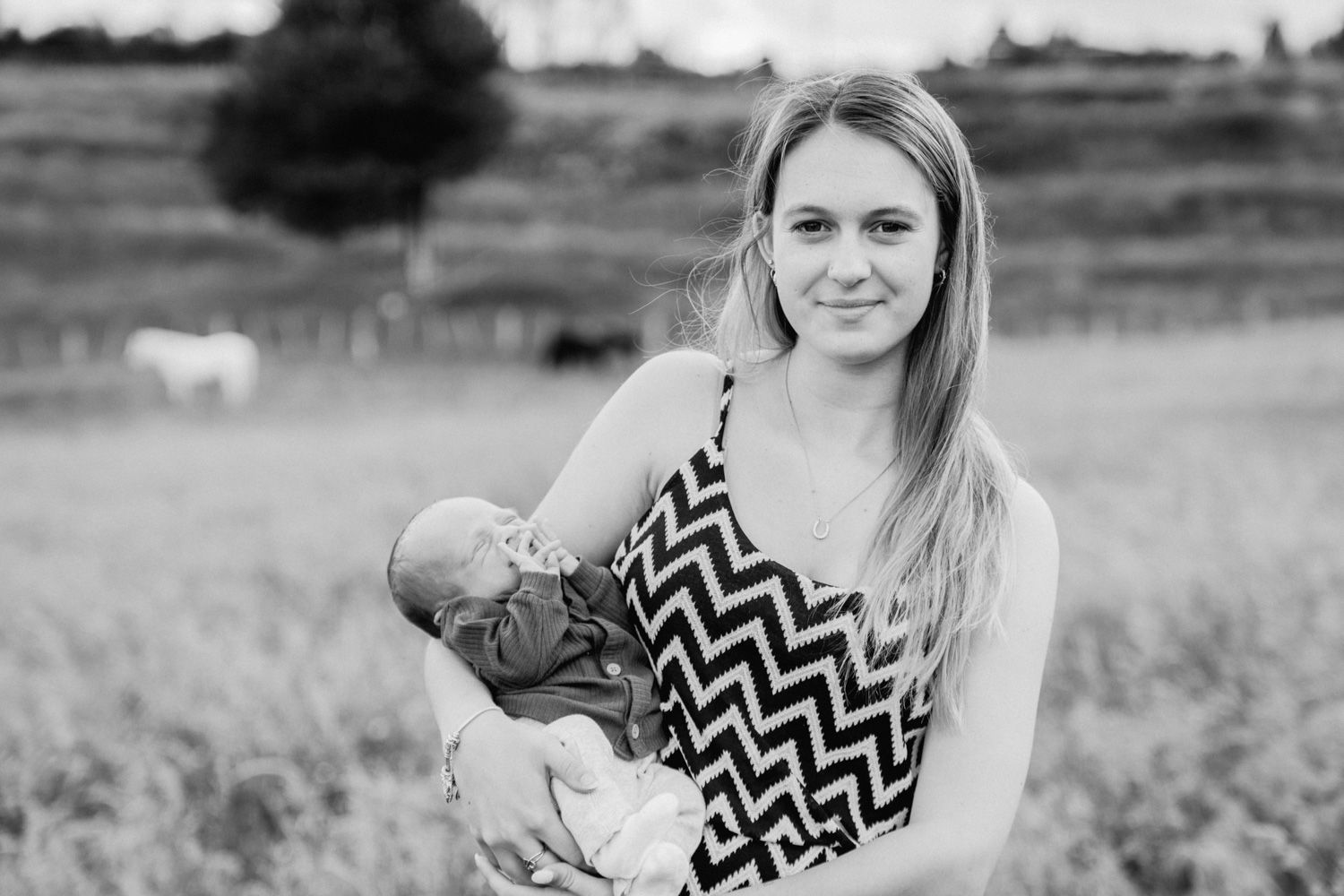 Black and white photo of a young woman holding a newborn baby in a field with a white horse in the background.