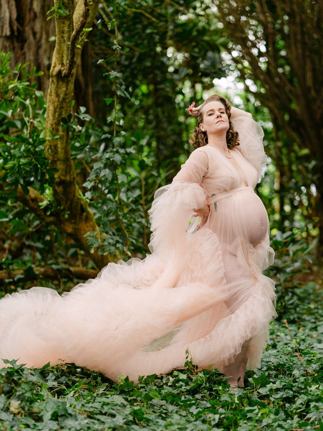 Pregnant woman in flowing blush pink tulle gown poses gracefully among lush green ivy and trees in an enchanting forest setting.