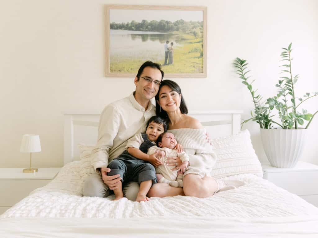 Happy family of four posing on a white bed, parents smiling with toddler and newborn baby in bright bedroom.