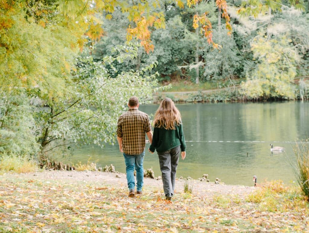 Couple walking hand in hand along a scenic autumn pond path surrounded by colorful fall foliage and geese.