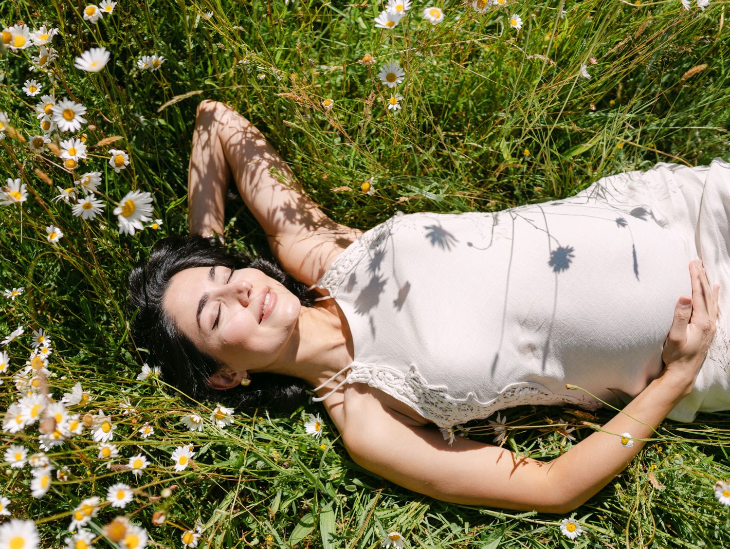A young woman in a floral dress lying peacefully in a sunny green meadow surrounded by white daisy wildflowers.