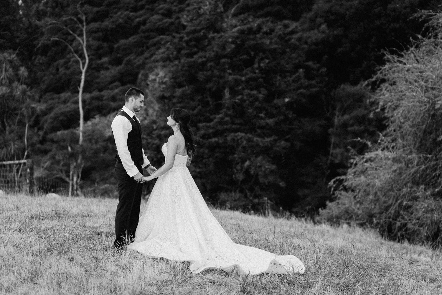 Black and white photo of a bride and groom sharing a romantic moment on a grassy hillside with lush forest backdrop.