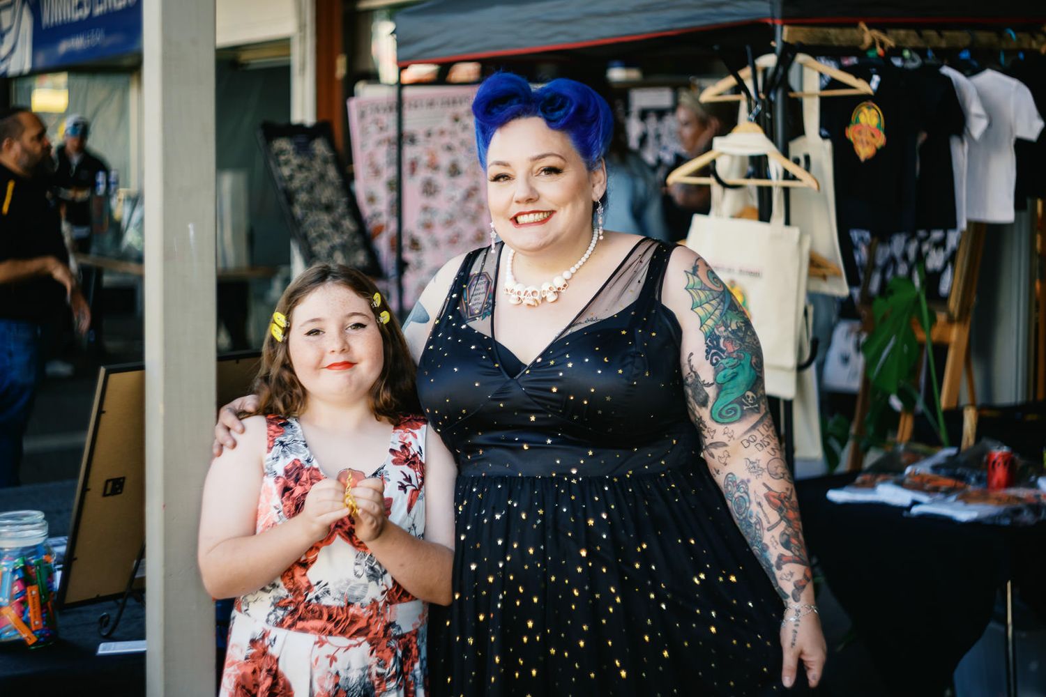 Two women smiling at an outdoor market, one in floral dress, one in black star dress with blue hair and tattoos.