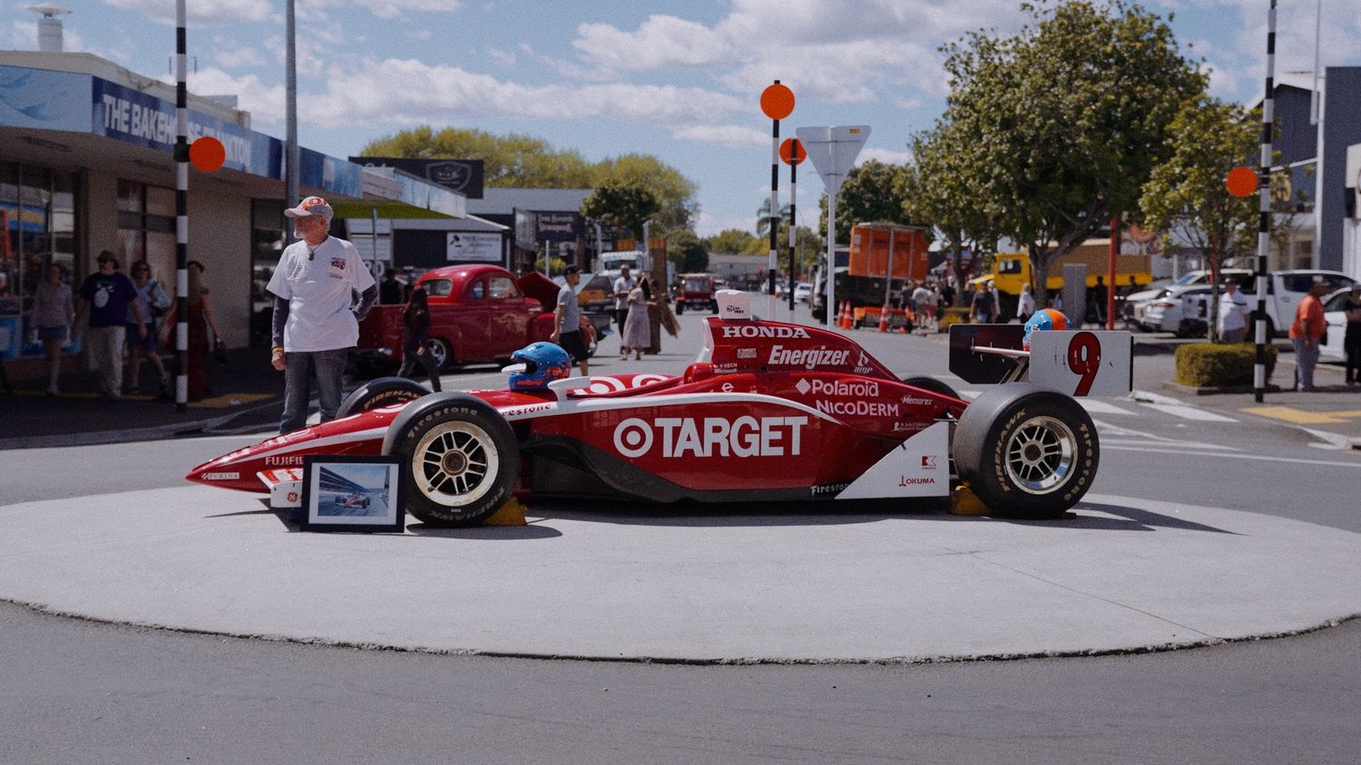 Red Target-sponsored IndyCar racing car on display in a town center roundabout with spectators and shops in background.