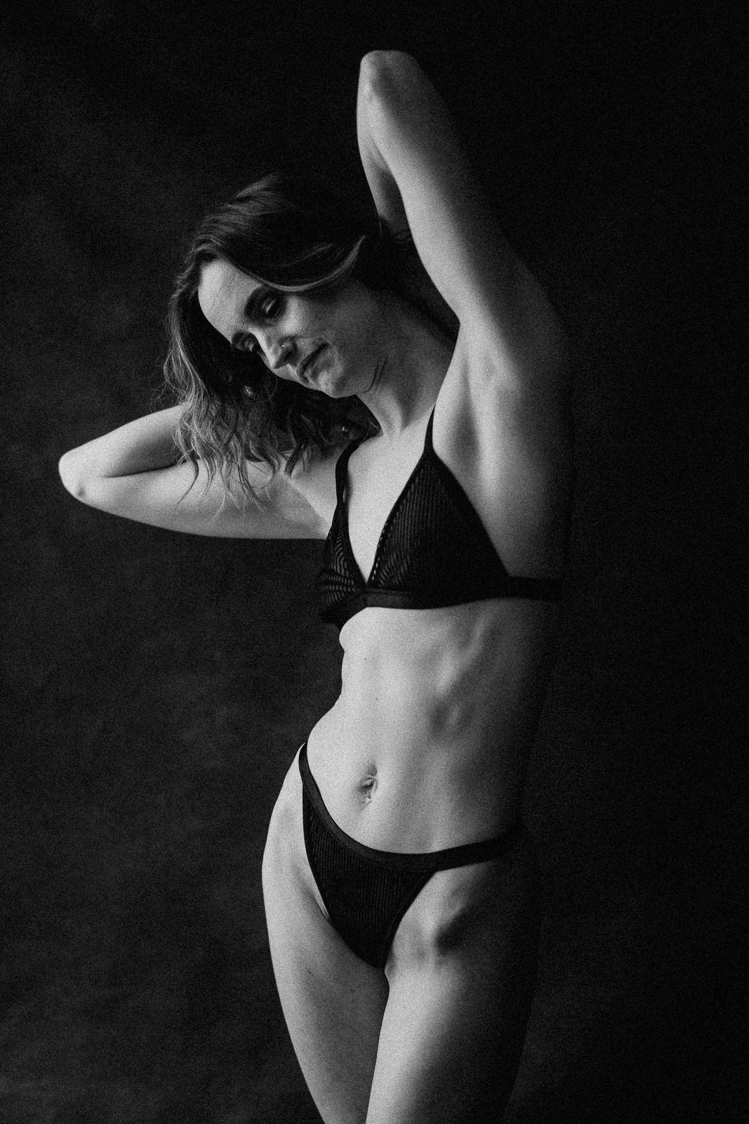 Black and white studio photo of a woman posing in a black bikini against a dark background.