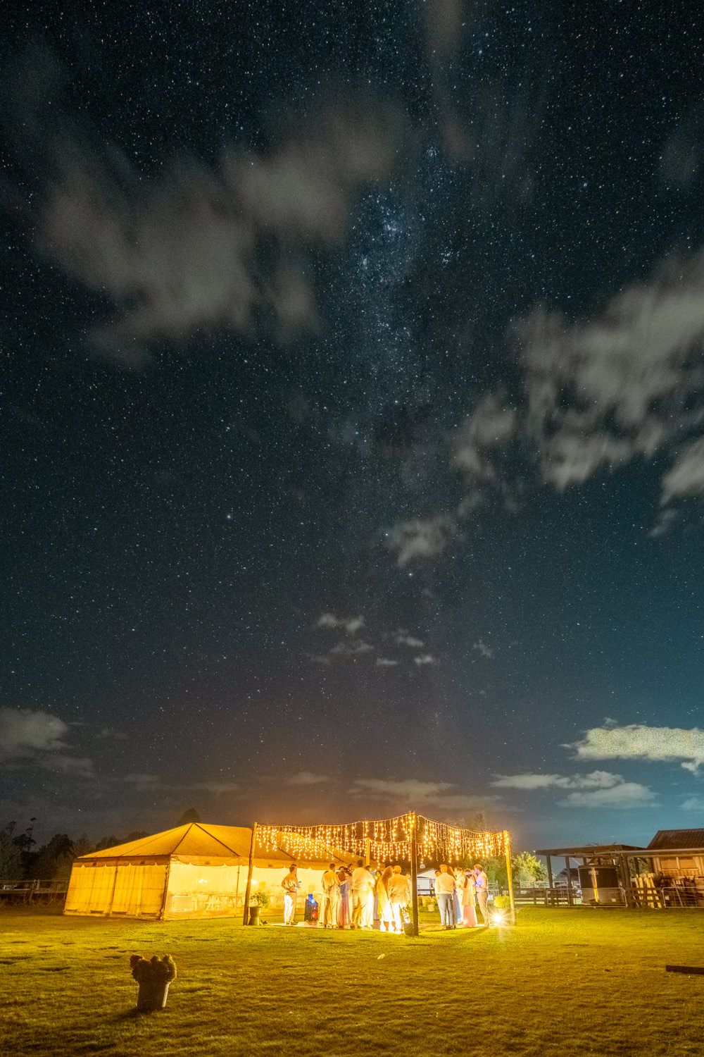 Starry night sky over illuminated wedding tent on green field with Milky Way visible through clouds.