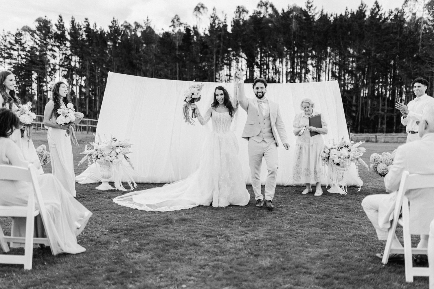 Black and white photo of newlyweds celebrating after outdoor wedding ceremony with guests and bridal party.