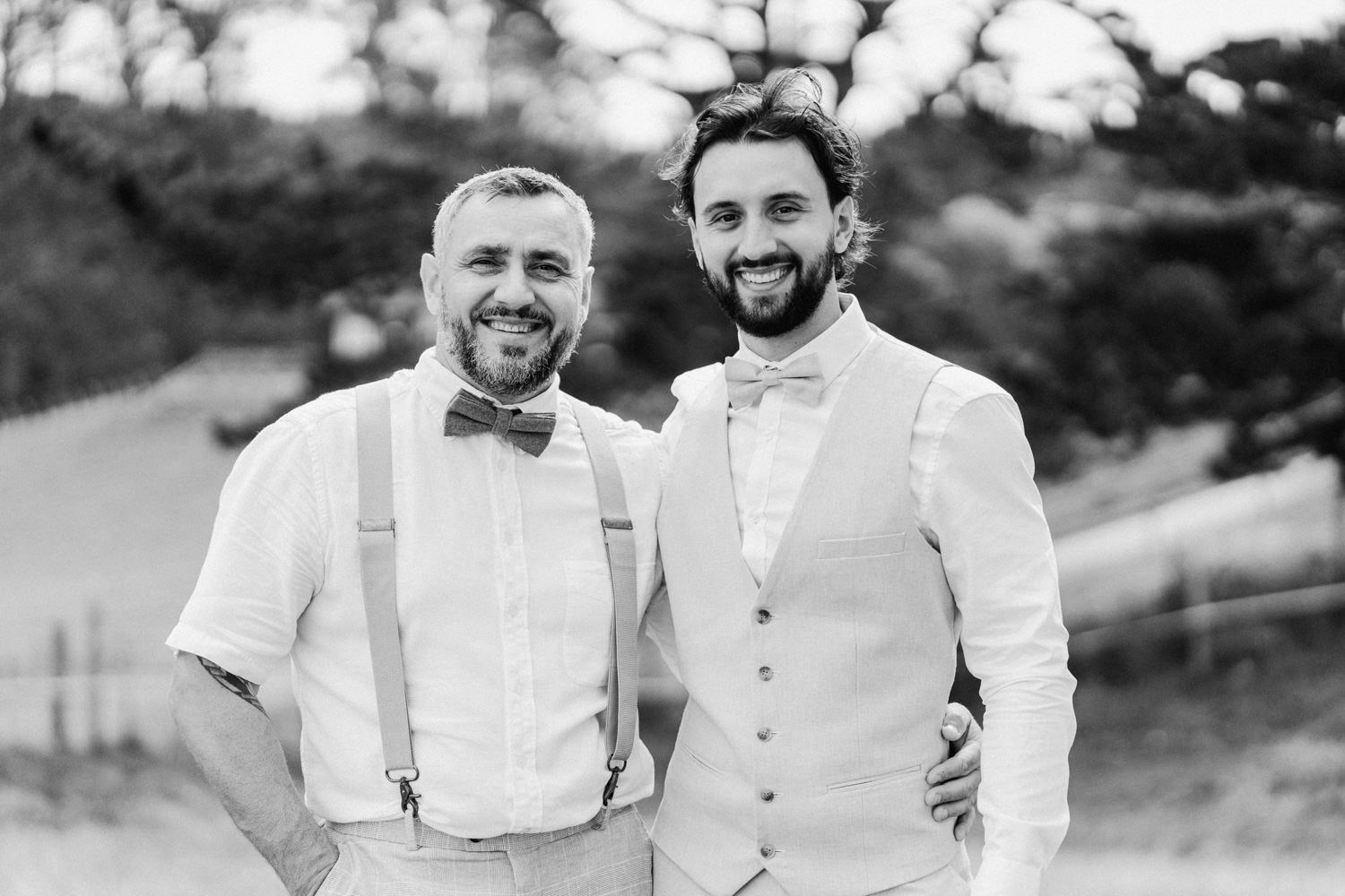 Two smiling men in bow ties and suspenders pose outdoors at a wedding celebration in black and white photo.