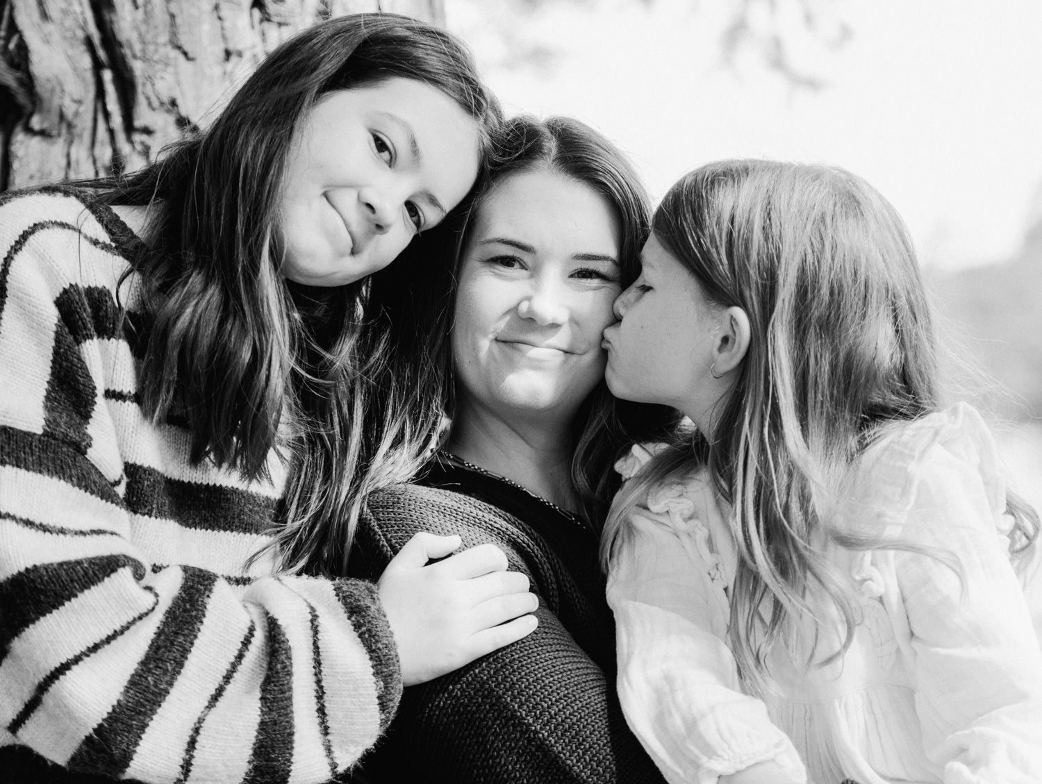 Black and white photo of a mother and two daughters sharing a tender embrace near a tree.