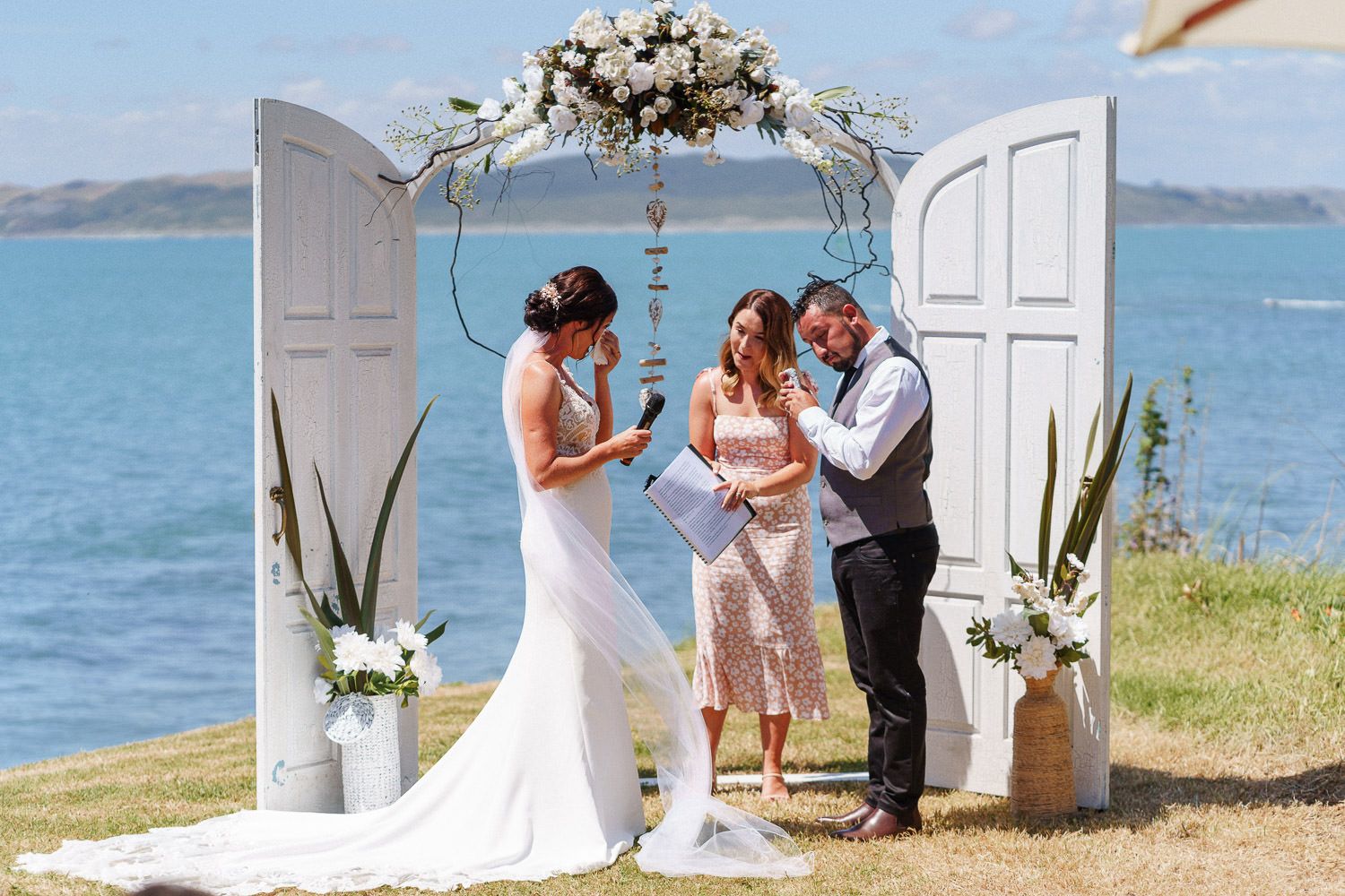 Couple exchanging vows at a waterfront wedding ceremony beneath a floral arch with white doors.