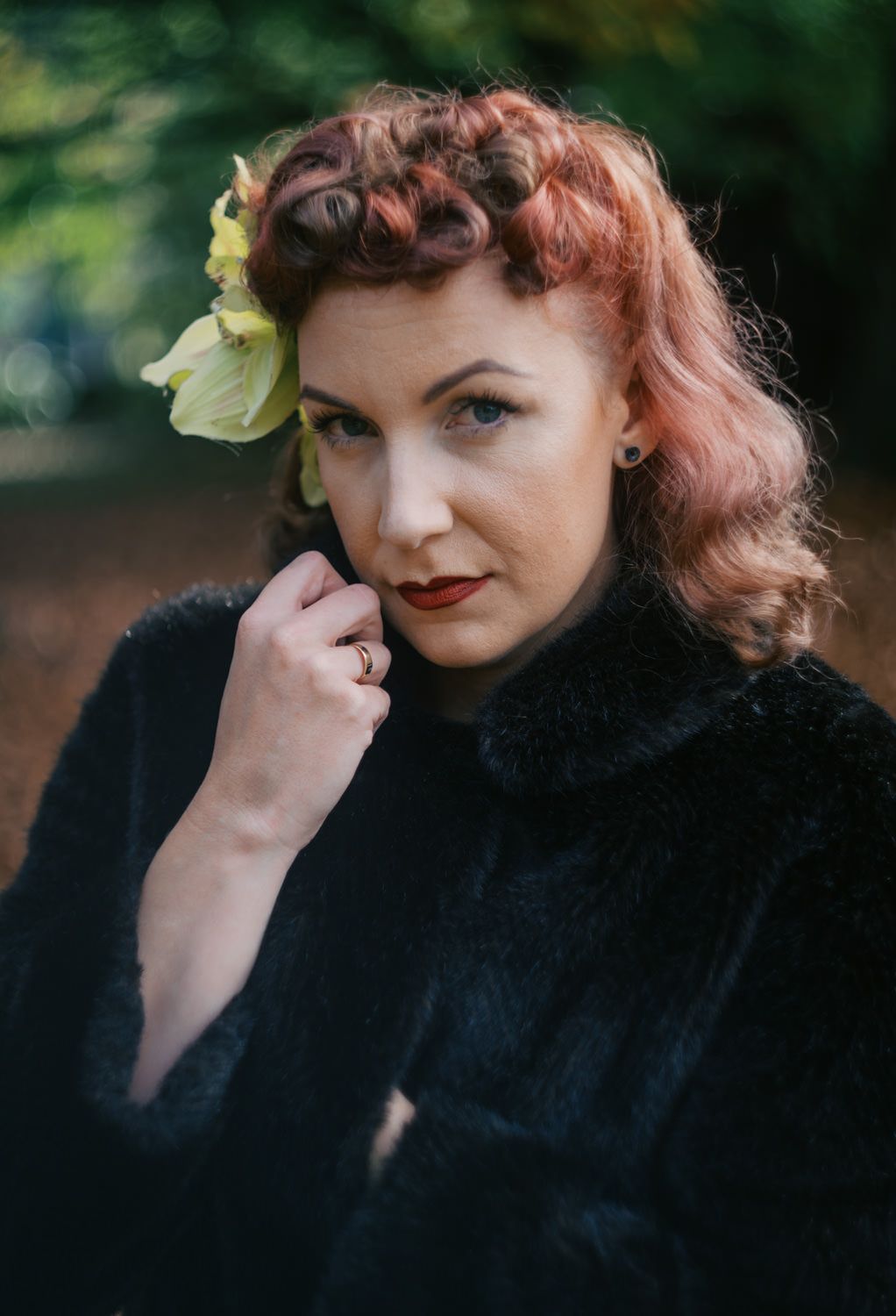 A woman with red hair and a yellow flower accessory poses in a black fur coat outdoors.