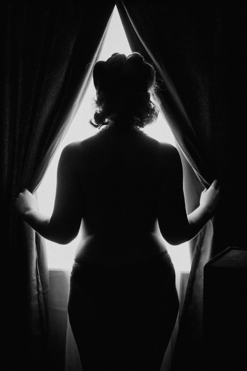 Black and white silhouette of a woman standing at a bright window, holding open curtains in a dramatic pose.
