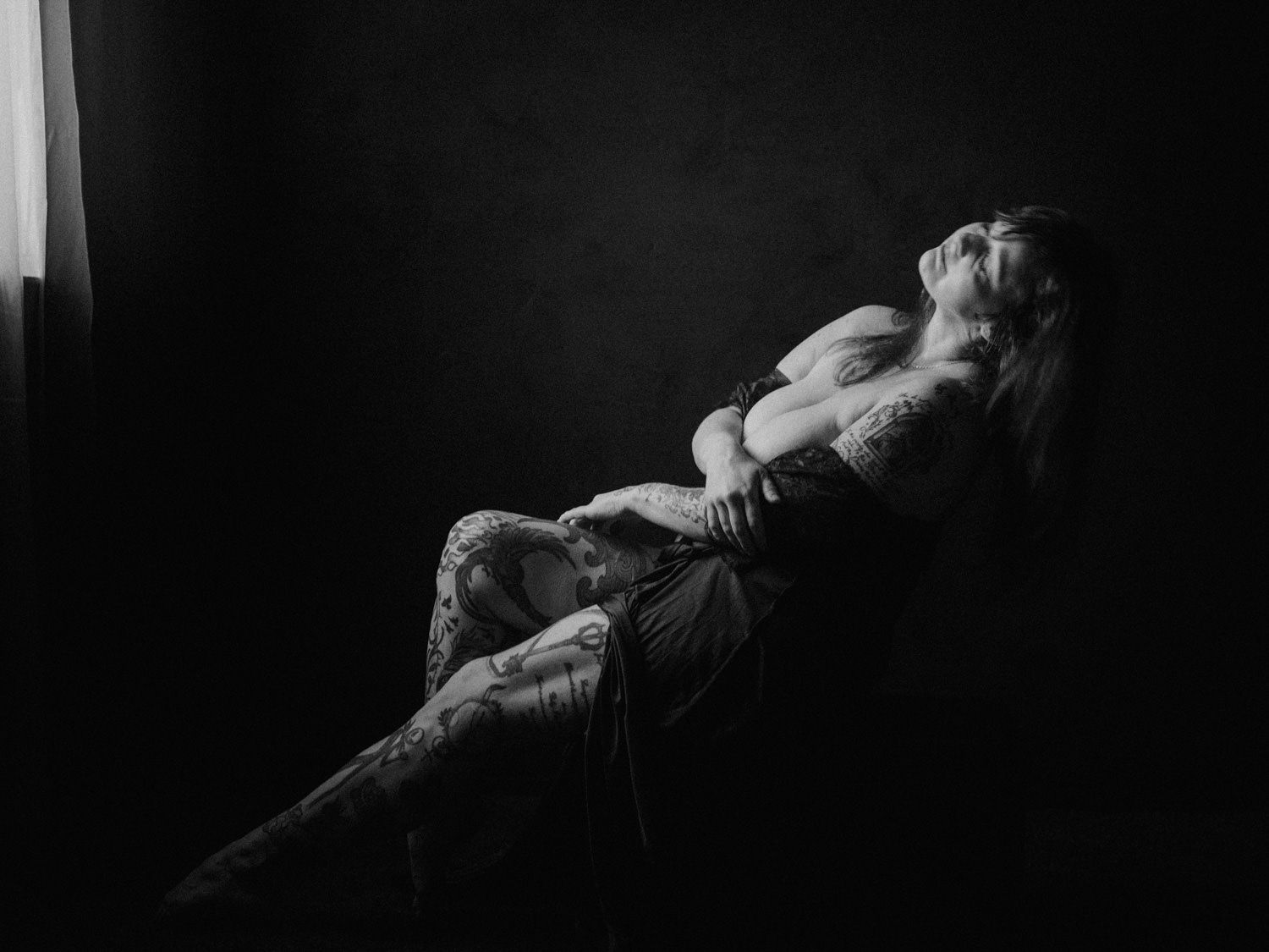 Black and white photo of a tattooed woman reclining dramatically in a chair near a window in moody lighting.