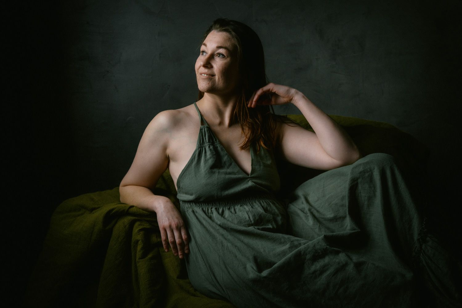 Woman in green dress poses gracefully on a green draped couch against a dark backdrop in a studio portrait.