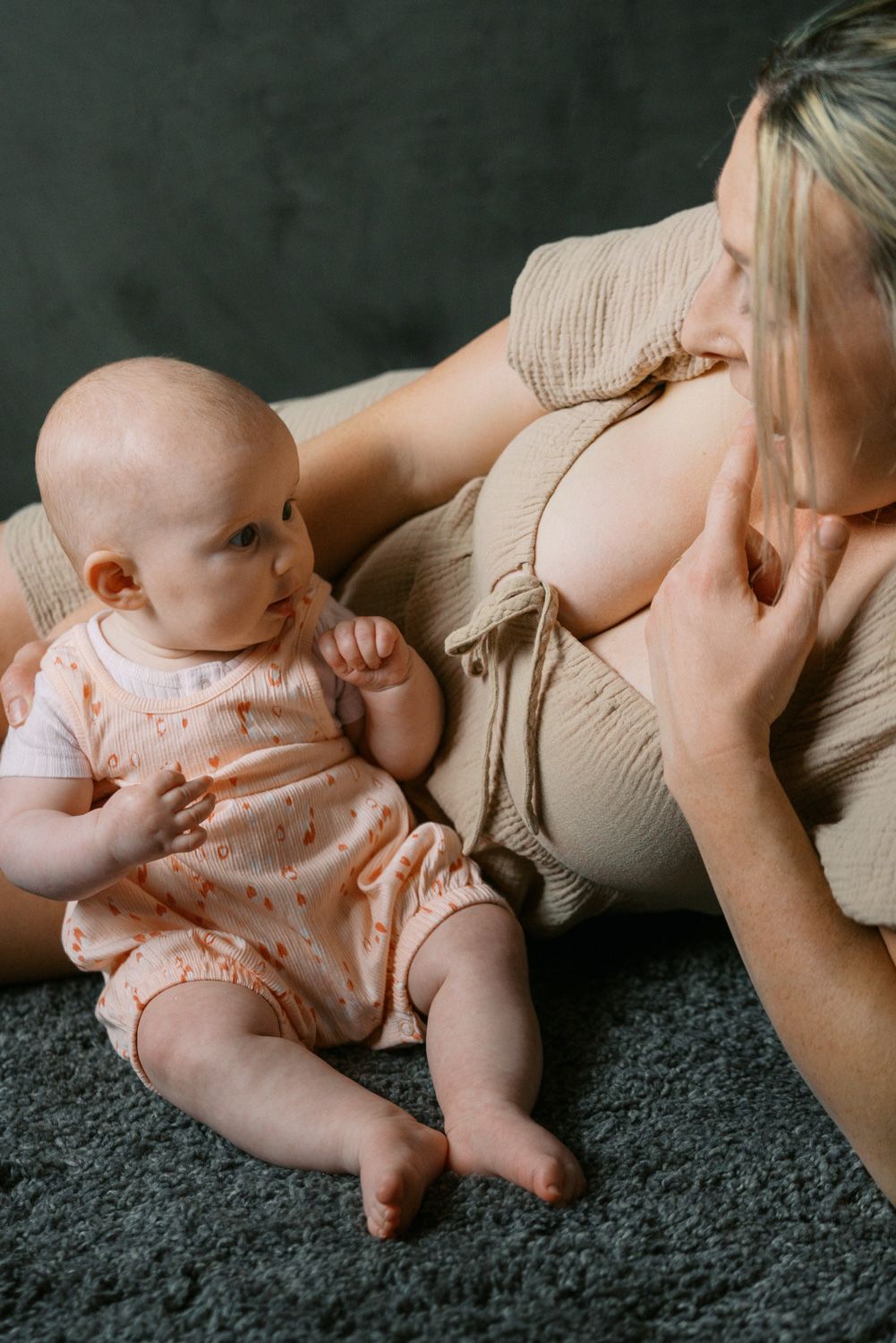 A baby in a pink romper sits on a gray carpet beside a blonde woman in a beige outfit.