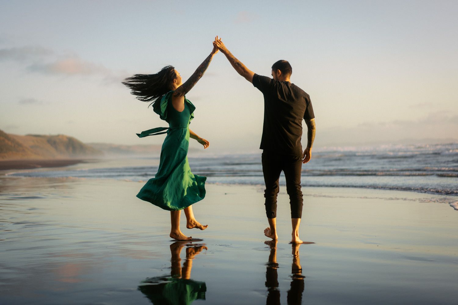 Couple dancing barefoot on a reflective beach at sunset, woman in green dress spinning with hair flowing in the wind.