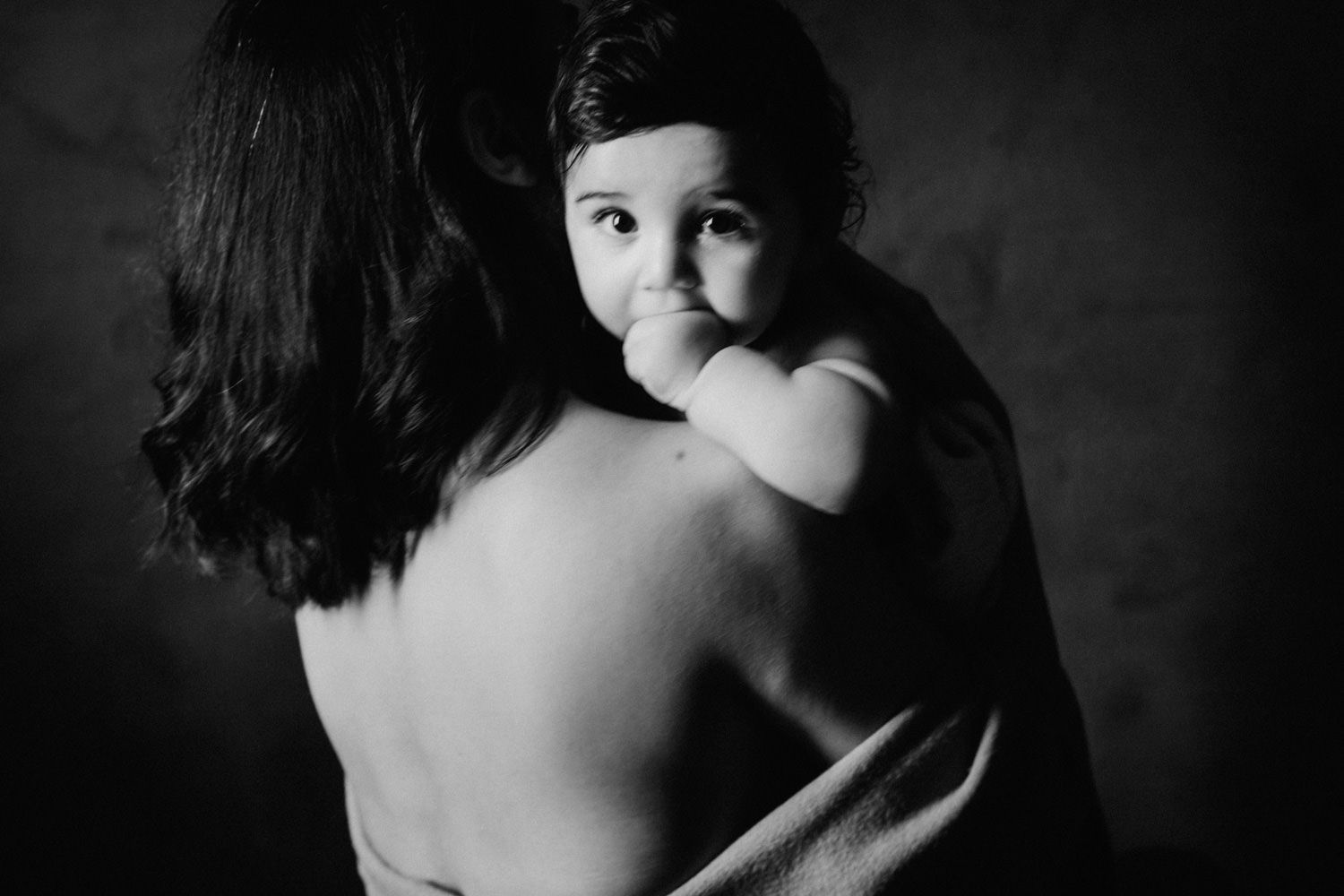 Black and white photo of a mother holding her baby close, the infant looking over her bare shoulder.