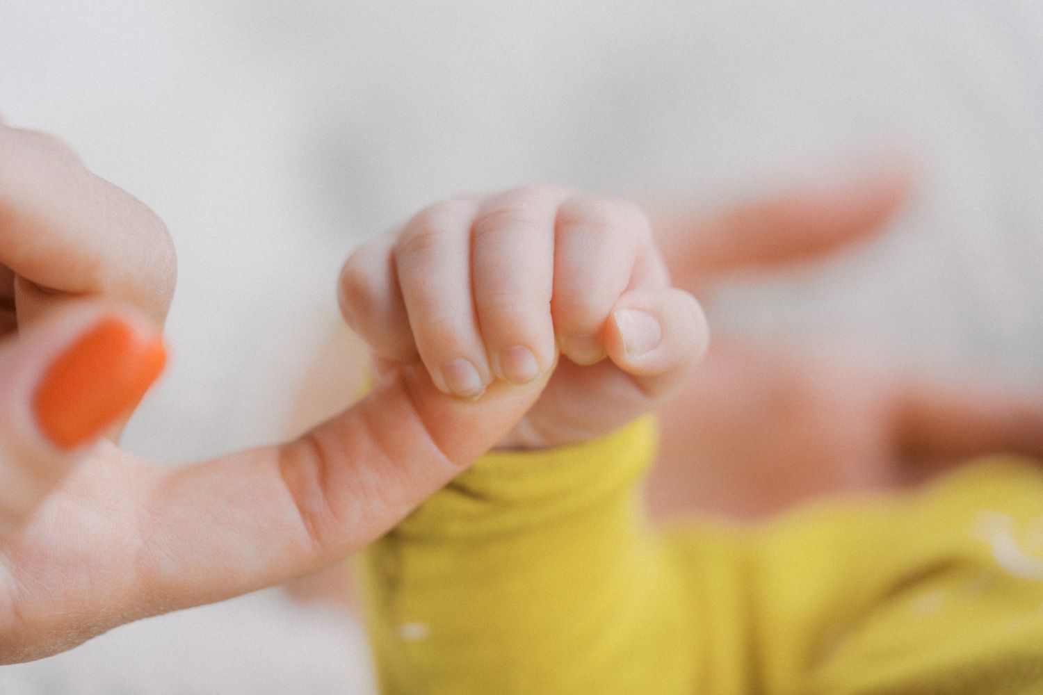 A baby grasping an adult's finger, wearing a yellow onesie, in a soft close-up shot.